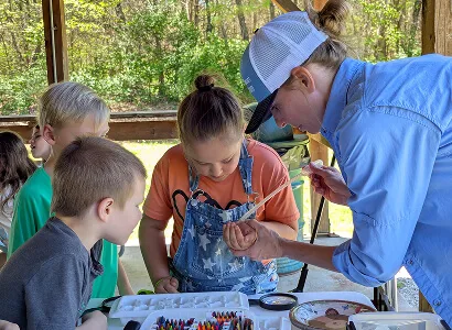 A woman helps two children closely examine an object with a dropper at an outdoor table covered with art supplies, crayons, and a palette, with trees visible in the background.
