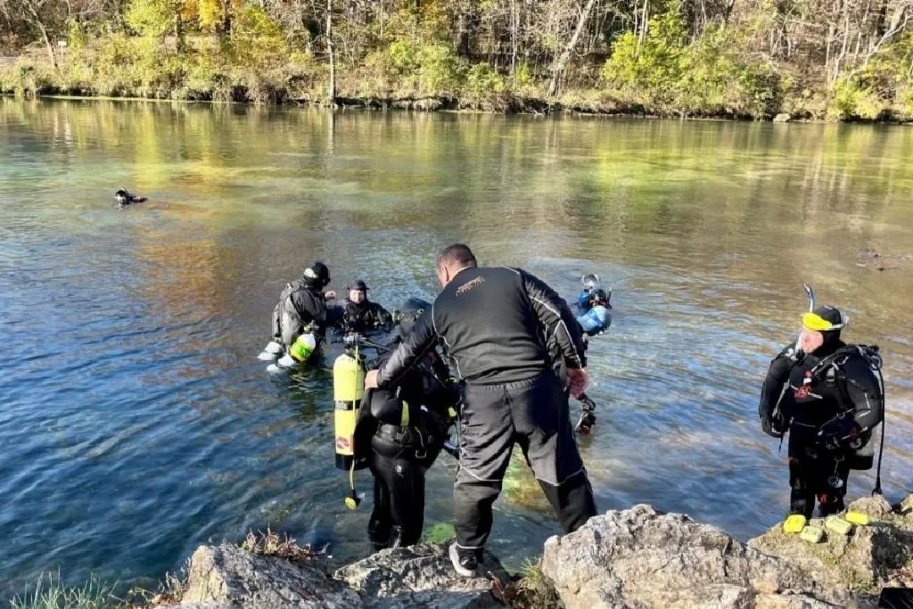 Several scuba divers in wetsuits are in a clear, shallow river near a rocky shore, preparing to enter or exit the water. Trees with autumn foliage are visible in the background.