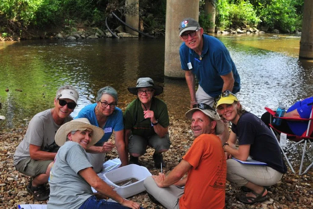 A group of six adults wearing hats sit and kneel on a rocky riverbank, gathered around containers and equipment, smiling at the camera. Trees and a bridge are visible in the background.