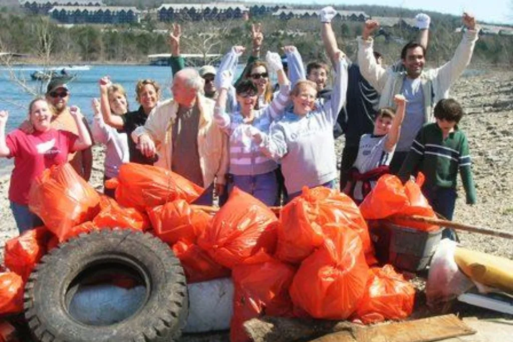 A group of smiling people cheer while standing behind a pile of collected trash in orange bags, tires, and debris on a rocky shoreline, with water and houses visible in the background.