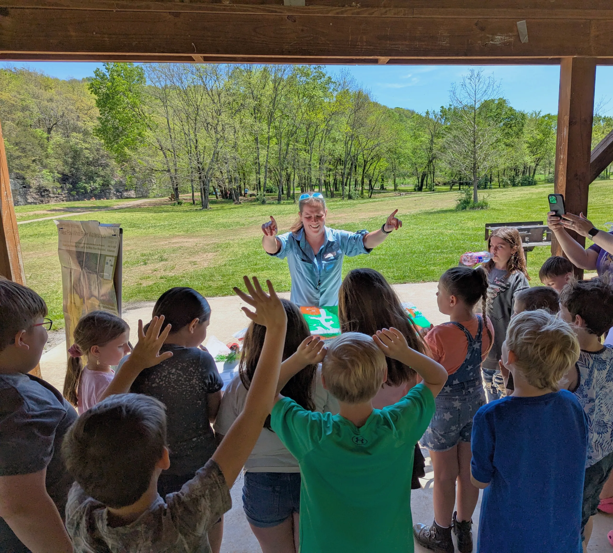 A group of children stand under a pavilion, enthusiastically raising their hands toward a smiling adult who is leading an outdoor educational activity. Trees and a grassy field are visible in the background.