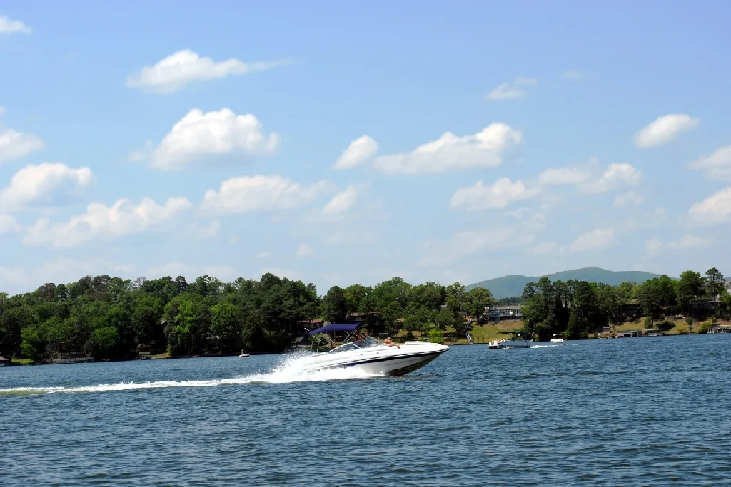 A white speedboat moves quickly across a blue lake, leaving a trail of white water. Trees and houses are visible along the shoreline under a partly cloudy sky.