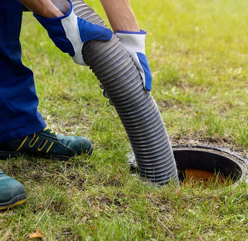 A person wearing gloves and work boots holds a large hose, inserting it into an open outdoor septic tank on a grassy lawn.