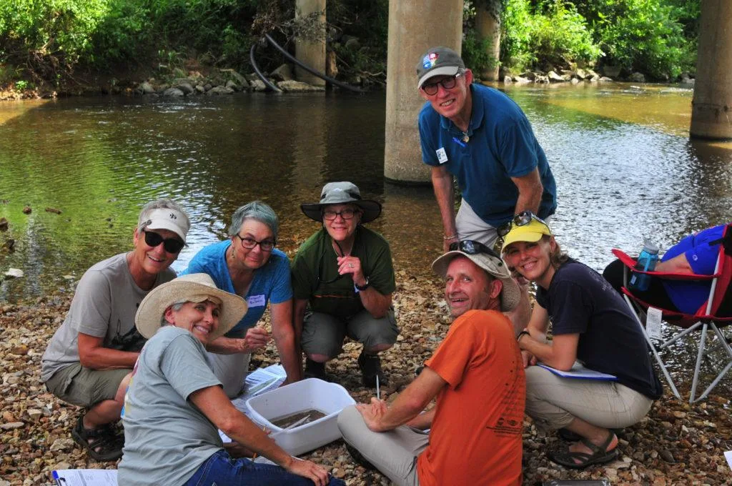 A group of seven adults, wearing hats and casual clothes, gather on a rocky riverbank by a shallow stream, smiling at the camera. They examine a white container with equipment, surrounded by greenery and bridge pillars.