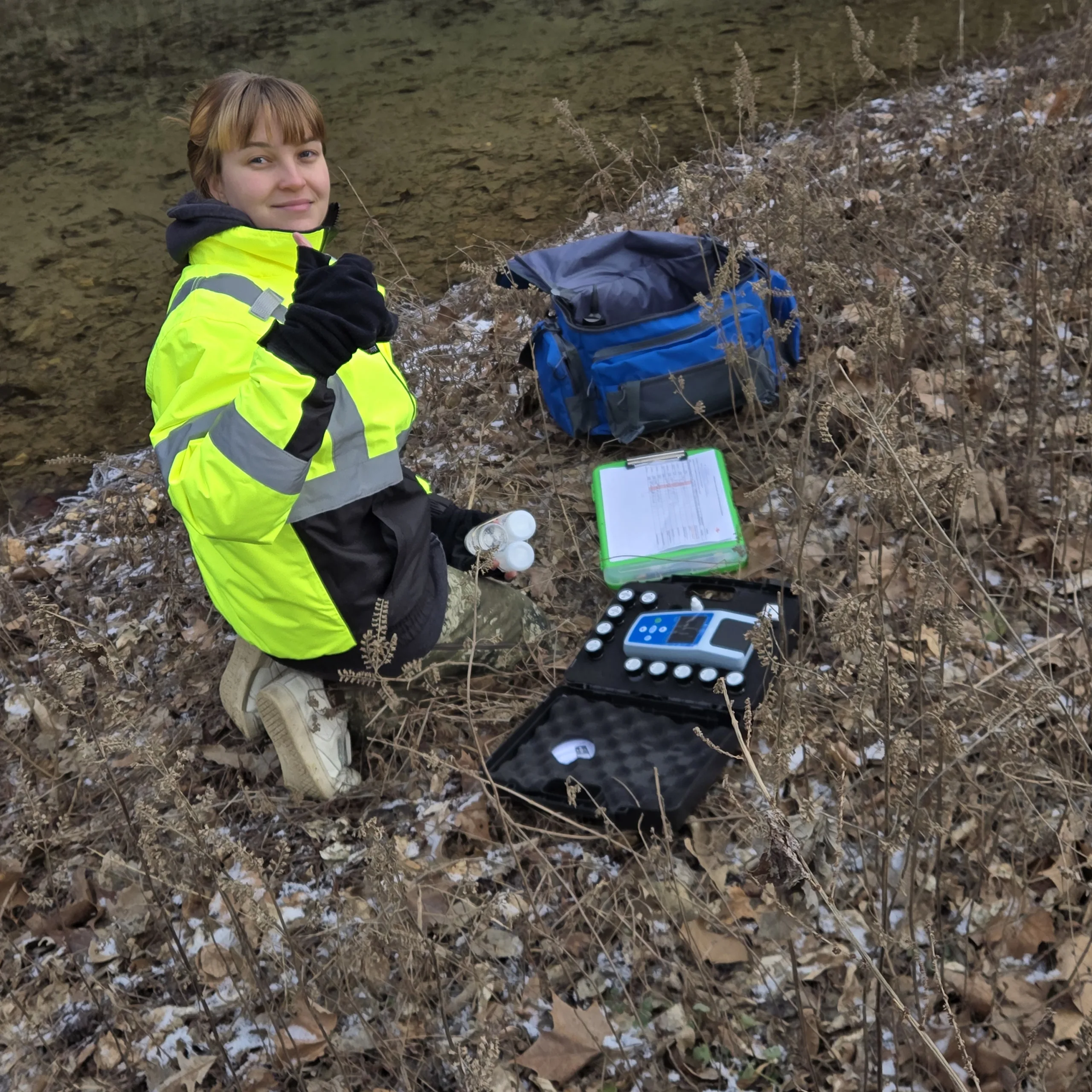 A person in a bright yellow reflective jacket kneels near a riverside, holding small bottles. Nearby are scientific equipment, a blue bag, and a clipboard, suggesting water sampling or environmental monitoring. Dry leaves cover the ground.